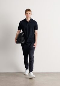 Joven caminando, viste una camiseta polo negra y pantalones, calza zapatillas blancas, lleva una gran bolsa negra bajo el brazo frente a un fondo blanco liso.