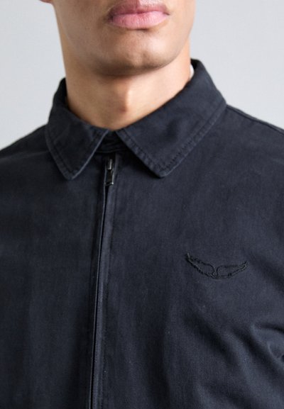 Black zip-up jacket with a collared neckline, featuring a textured fabric and stitched wing logo detail on the chest.