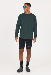 Langärmliges, dunkelgrünes Sportshirt mit Raglanärmeln, schwarze Kompressionsshorts, schwarze Socken und schwarze Radschuhe. Blaue Sonnenbrille.