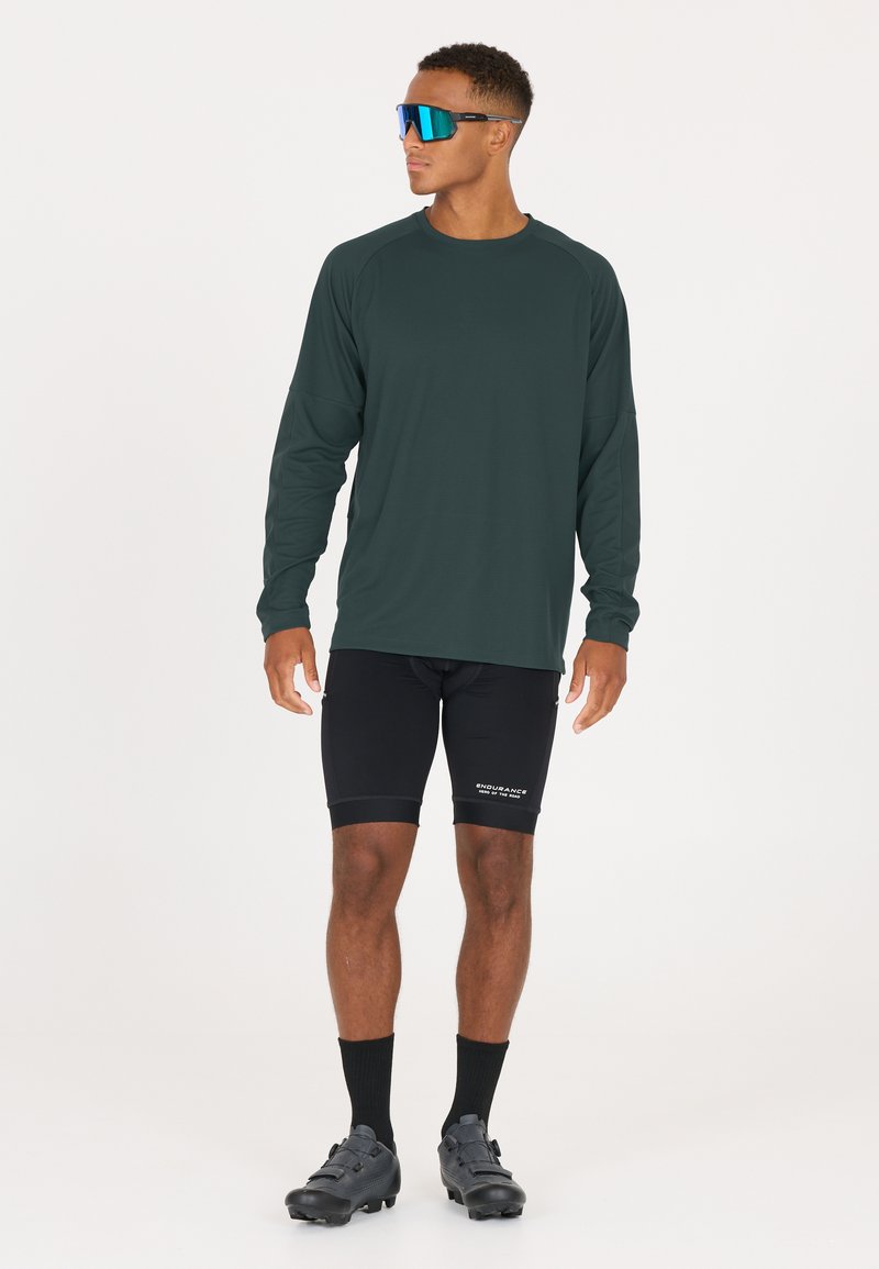 Langärmliges, dunkelgrünes Sportshirt mit Raglanärmeln, schwarze Kompressionsshorts, schwarze Socken und schwarze Radschuhe. Blaue Sonnenbrille.