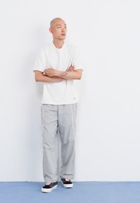Camicia bianca a maniche corte, pantaloni grigi larghi con strisce bianche sui lati e scarpe nere con lacci rosa. In piedi davanti a uno sfondo bianco.