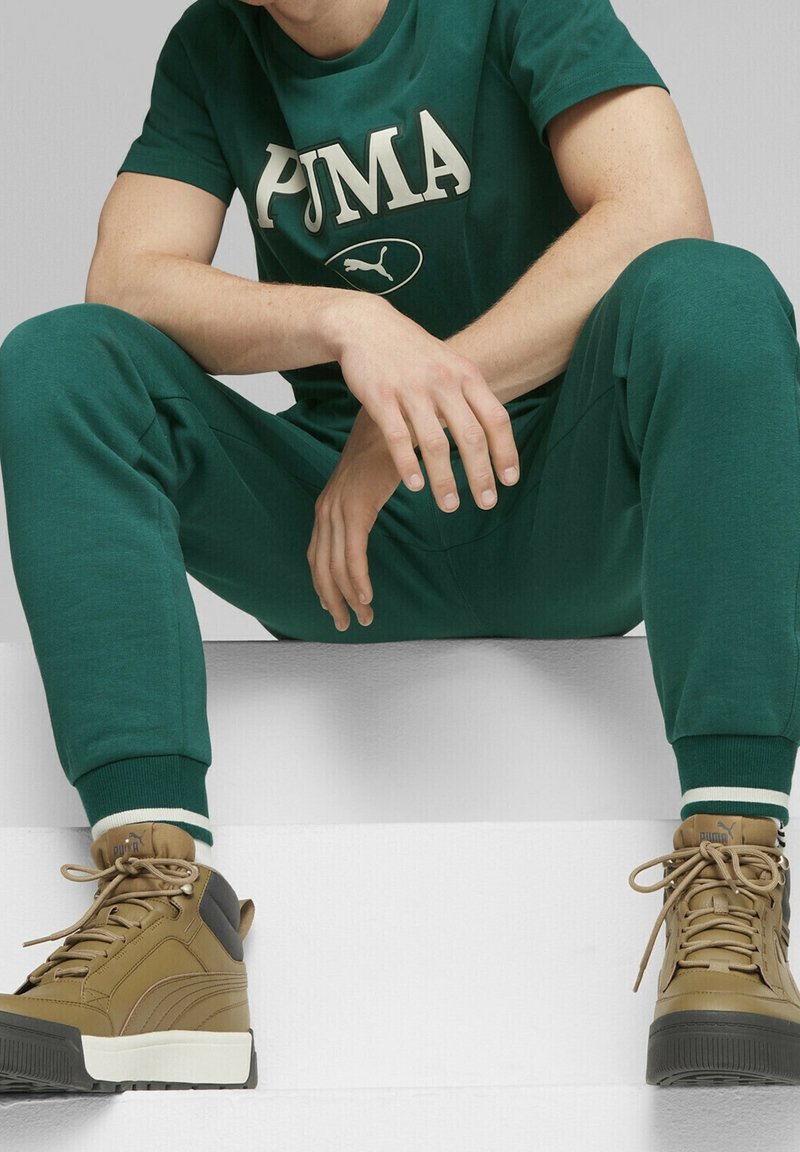 Grön bomullströja med "PUMA"-logotyp, i kombination med matchande joggerbyxor. Bruna högskurna skor med vit sula, med snörningsdesign.