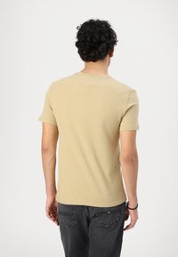 T-shirt corto color sabbia con collo rotondo, realizzato in tessuto morbido. Il retro presenta un yoke a forma di V. Abbinato a jeans grigio scuro.