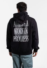Felpa nera in cotone con vestibilità rilassata, caratterizzata da una grande scritta grafica bianca sul retro che recita "SEMPLICEMENTE SEMPRE UN LAVORO IN CORSO."