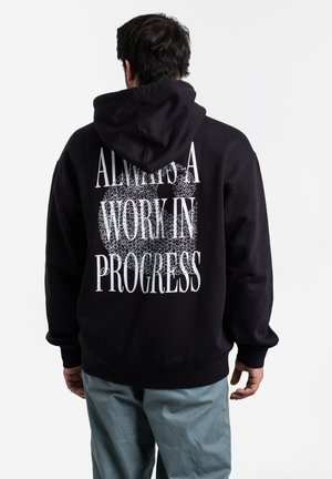 Black hoodie made of cotton with a relaxed fit, featuring a large white graphic text design on the back that reads "ALWAYS A WORK IN PROGRESS."