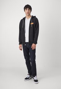 Sudadera con capucha negra y cremallera frontal, con un logo en naranja, combinada con una camiseta blanca, jeans oscuros y zapatillas negras.
