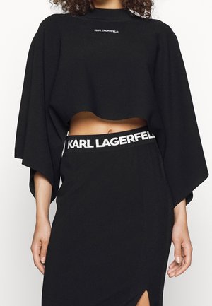 Frau trägt ein schwarzes, kurzes Oberteil mit weiten Ärmeln und einen hoch taillierten Rock mit dem "Karl Lagerfeld"-Logo am Ausschnitt und Bund.