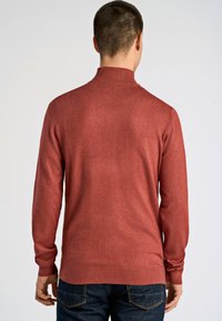 Pull rouge en tricot avec un col haut, des manches longues et des poignets côtelés. Tissu texturé avec une finition lisse. Porté sur un jean foncé.