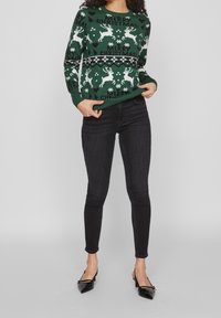 Grüner Strickpullover mit weißen Rentier- und Schneeflockenmuster, versehen mit dem Text "FROHE WEIHNACHTEN", kombiniert mit schwarzen Skinny-Jeans und spitzen Schuhen.