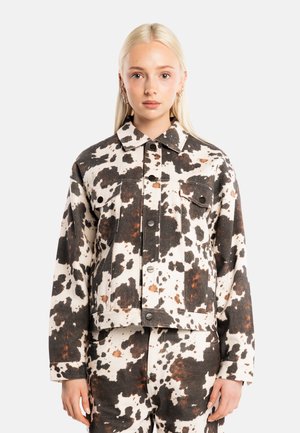 Joven mujer con cabello rubio largo que lleva una chaqueta y pantalones a juego con estampado de vaca en blanco y negro, de pie contra un fondo liso.