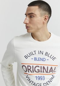 Felpa chiara con scollo rotondo. Grafica testuale in blu, arancione e marrone scuro che recita "Built in Blue, Blend, Original 1993, Vintage Denim."