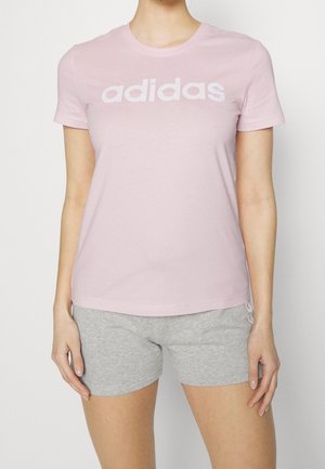 Kobieta w jasnoróżowej koszulce Adidas z białym logo i jasnoszarych szortach, stojąca na jednolitym białym tle.
