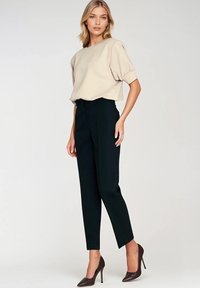 Beżowa bluzka z krótkim rękawem i okrągłym dekoltem, zestawiona z dopasowanymi czarnymi spodniami oraz brązowymi czółenkami na szpilce, na tle neutralnym.