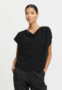 Top nero a maniche corte con scollo drappeggiato e texture morbida. Il tessuto ha una leggera brillantezza, abbinato a pantaloni neri su misura.
