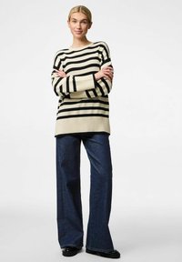 Sweter w paski w odcieniach kremu i czerni, z prążkowanymi mankietami, w zestawieniu z szerokimi spodniami dżinsowymi w ciemnym kolorze. Modelka stoi z założonymi rękami.