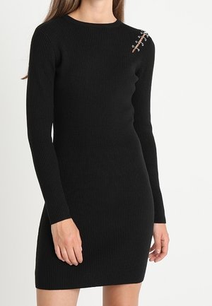 Robe longue à manches longues en maille côtelée noire avec un détail décoratif d'épingle de sûreté sur l'épaule gauche, portée par une femme.