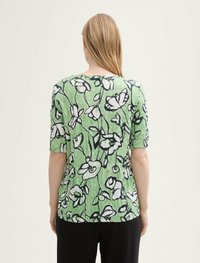 Grünes Oberteil mit floralem Muster und kurzen Ärmeln, das weiße Blumen zeigt, die in Schwarz umrandet sind, sowie einem strukturierten, zerknitterten Stoff.