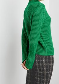 Vihreä neulottu korkeakauluksinen paita, jossa on ribbineulosta ja pitkät hihat, yhdistetty mustavalkoisiin ruutukuosiin housuihin.