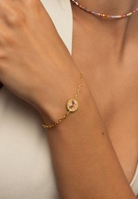 Bracciale d'oro con un ciondolo rotondo decorato da una farfalla viola, con catena a maglie e superficie texture, appoggiato su un polso abbronzato.