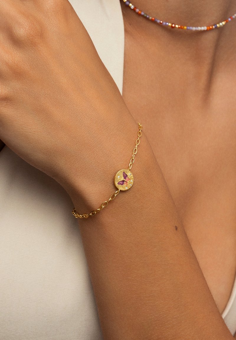 Bracciale d'oro con un ciondolo rotondo decorato da una farfalla viola, con catena a maglie e superficie texture, appoggiato su un polso abbronzato.