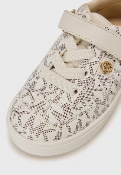 Zapatilla blanca con una superficie texturizada que presenta un patrón geométrico en marrón oscuro. Tiene un diseño de cordones en la parte frontal y una amplia correa que cruza el empeine.