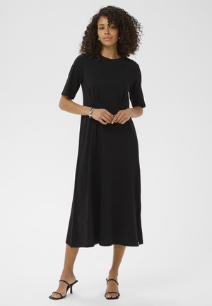 Femme aux cheveux bouclés portant une robe midi noire à manches courtes et des sandales noires à talons, debout devant un fond uni.