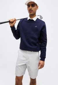 Άνδρας που φοράει λευκό καπέλο του γκολφ, μπλε πουλόβερ με το λογότυπο "ck", λευκό πουκάμισο με γιακά και ανοιχτό γκρι σορτς, κρατώντας μπαστούνι του γκολφ στον ώμο.