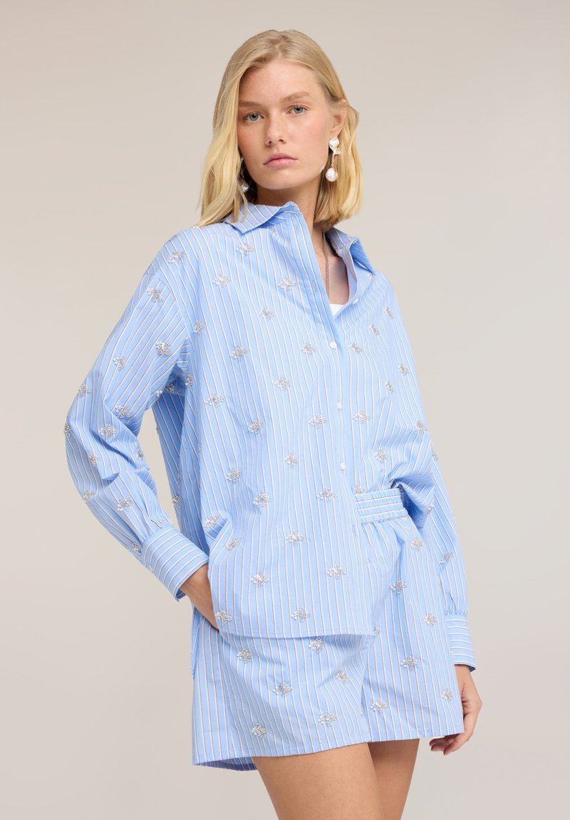 Mujer rubia con camisa azul claro a rayas finas y falda a juego con pequeño bordado floral, posando con una mano en el bolsillo.