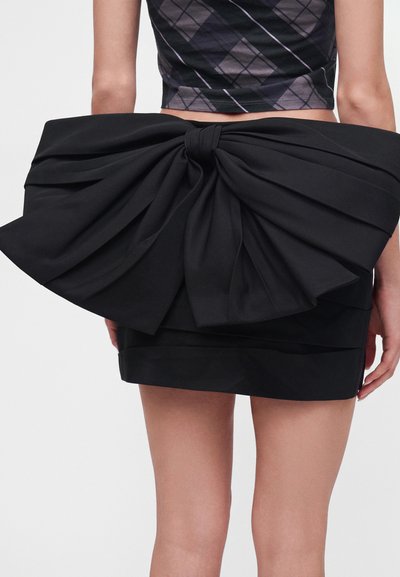 Femme portant une mini-jupe noire avec un grand nœud en tissu noir oversize noué dans le dos et un crop top sans manches à motif losanges.