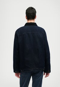 Tamsiai mėlynas džinsinis švarkas su laisvu siluetu, smaigiu apykakle, matomomis siūlėmis ir užsegimo detalėmis ant rankogalių.