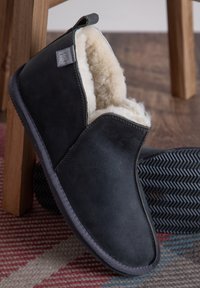Black suede slipper with faux fur lining, slip-on design, gray sole, and textured rubber outsole. Stitched detailing and brand tag visible.