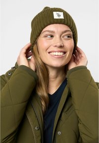 Berretto a coste verde oliva con una patch quadrata, indossato con una giacca olive con zip. Chi lo indossa ha i capelli lunghi e un'espressione sorridente.