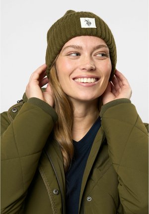 Berretto a coste verde oliva con una patch quadrata, indossato con una giacca olive con zip. Chi lo indossa ha i capelli lunghi e un'espressione sorridente.