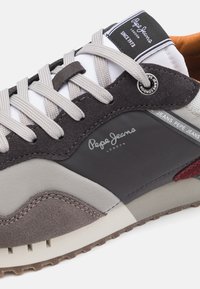 Gray and black sneaker with textured suede and smooth fabric, featuring gray laces, a white tongue, and a logo detail on the side.