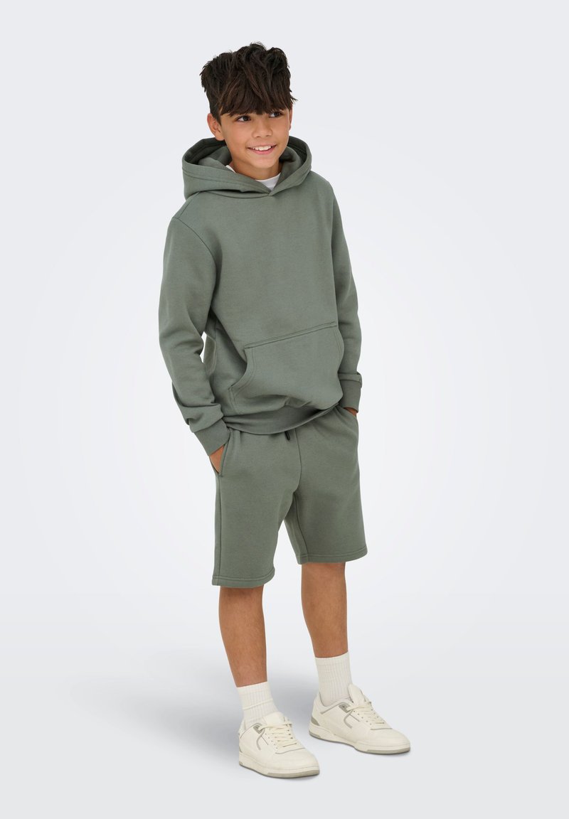 Grøn bomulds hoodie med frontlomme, parret med matchende shorts. Høje hvide sneakers og ribbede crew-sokker fuldender outfittet.