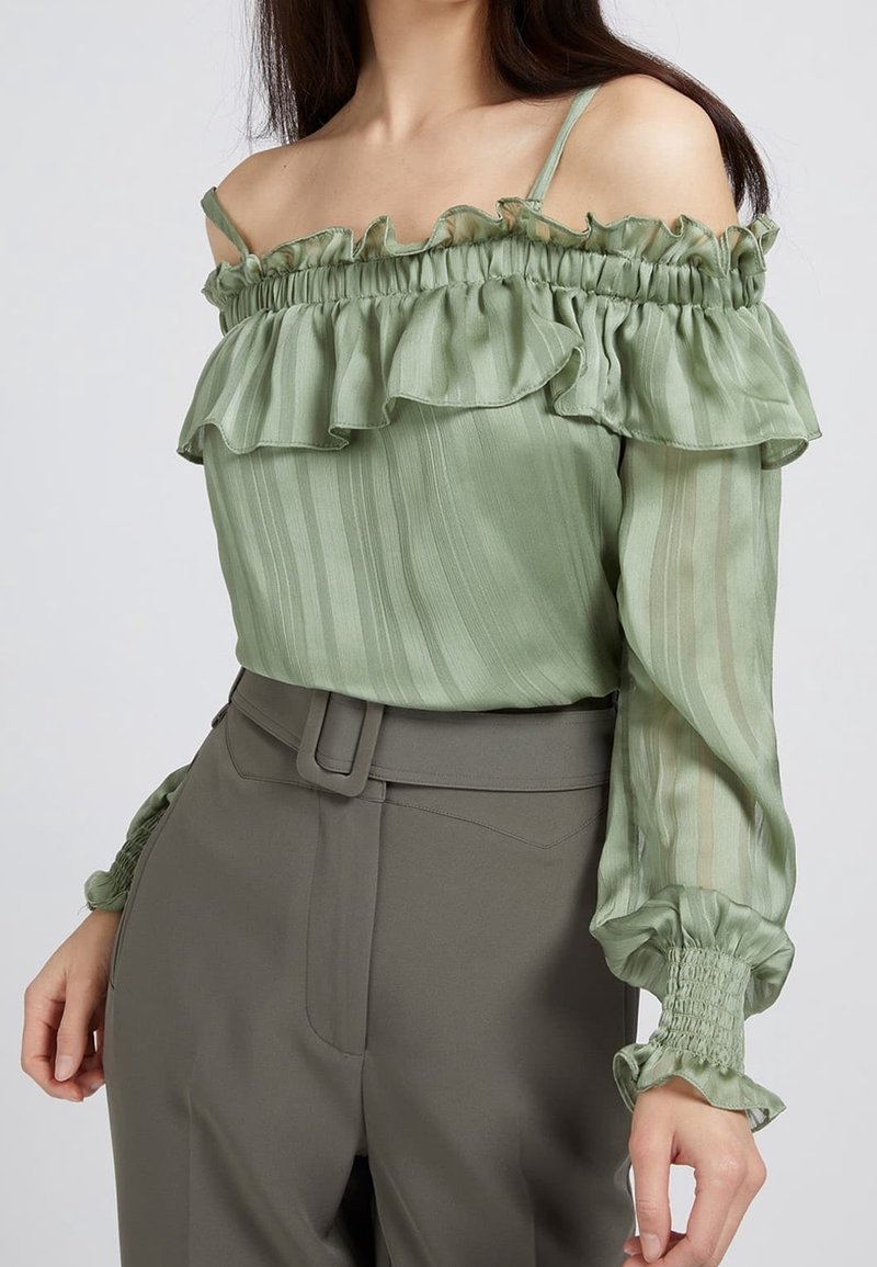 Blusa off-shoulder in verde chiaro, con bordo a volant, tessuto trasparente a righe e polsini arricciati. Abbinata a pantaloni in olive a vita alta.