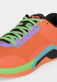 Φωτεινό πορτοκαλί αθλητικό παπούτσι με πράσινα κορδόνια, μωβ λεπτομέρειες και πράσινο-μπλε σόλα σε λευκό φόντο.