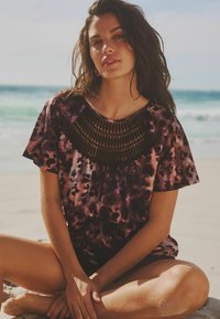 Krátký rukáv v purpurovo-růžovém tie-dye vzoru s texturovaným černým háčkovaným detailem kolem výstřihu, nošený osobou sedící na písku.
