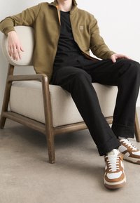 Person som bär brun jacka, svart skjorta, svarta byxor och bruna-vita sneakers, sittande på en beiga vadderad trästol på betonggolv.