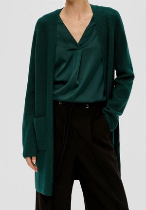 Cardigan en maille teal avec une ouverture à l'avant, des manches côtelées et une poche frontale, associé à un blouse vert foncé et un pantalon noir.