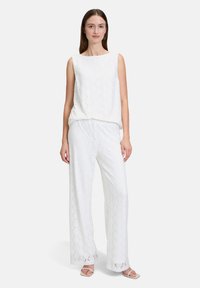 Biała bezrękawnik i pasujące szerokie spodnie wykonane z fakturowanej tkaniny w diamentowy wzór, zestawione z jasnymi sandałami.