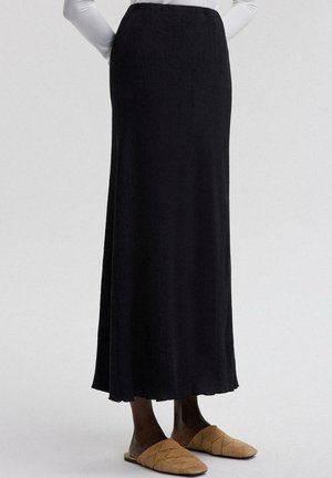 Kvinde iført en lang sort nederdel med tekstureret stof og tan farvede quiltede slip-on sko, stående med hænderne bag ryggen.