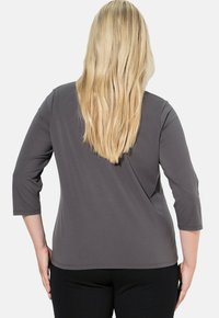 Vaaleatukkainen nainen, jolla on pitkät hiukset, pukeutunut tummanharmaaseen 3/4 hihaiseen toppiin ja mustiin housuihin, kuvattuna takaa valkoista taustaa vasten.