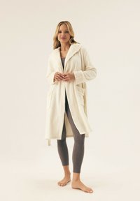 Kremowy szlafrok z polaru z kapturem, prążkowanymi mankietami i bocznymi kieszeniami. Noszony na szarych legginsach, ukazujący miękką, teksturowaną tkaninę.