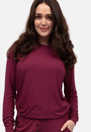 Top lungo bordeaux con maniche lunghe realizzato in un tessuto morbido, caratterizzato da una vestibilità comoda, scollatura a barca e leggero arricciamento in vita. Dettagli con bottoni neri.