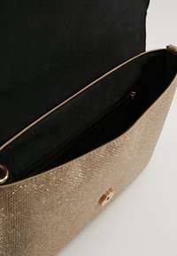 Beżowa torebka kopertowa z brokatem o gładkiej fakturze, złotym okuciem i zapinana na zamek wewnętrzną kieszonką. Wnętrze wyłożone jest czarną tkaniną.