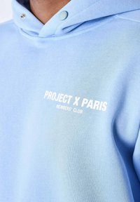 Sudadera azul claro hecha de tejido de mezcla de algodón, con capucha ajustable, botones a presión y texto blanco: "CLUB DE MIEMBROS DE PROJECT X PARIS."