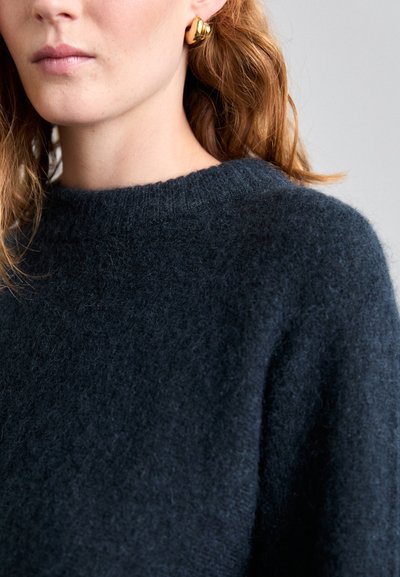 Pull en laine bleu marine avec un col rond et une texture douce, associé à des boucles d'oreilles en or pour une touche supplémentaire.