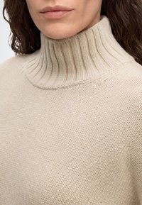 Femme portant un pull à col roulé en maille de couleur crème avec un col côtelé et un tissu texturé ample.