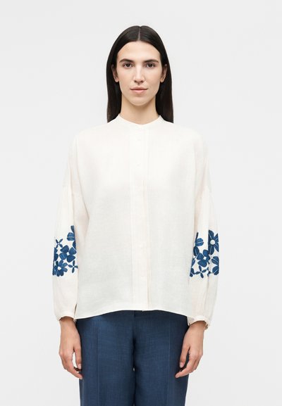 Femme aux cheveux lisses et foncés porte un chemisier blanc à manches longues avec une broderie florale bleue sur les manches et un pantalon bleu, debout devant un fond blanc.